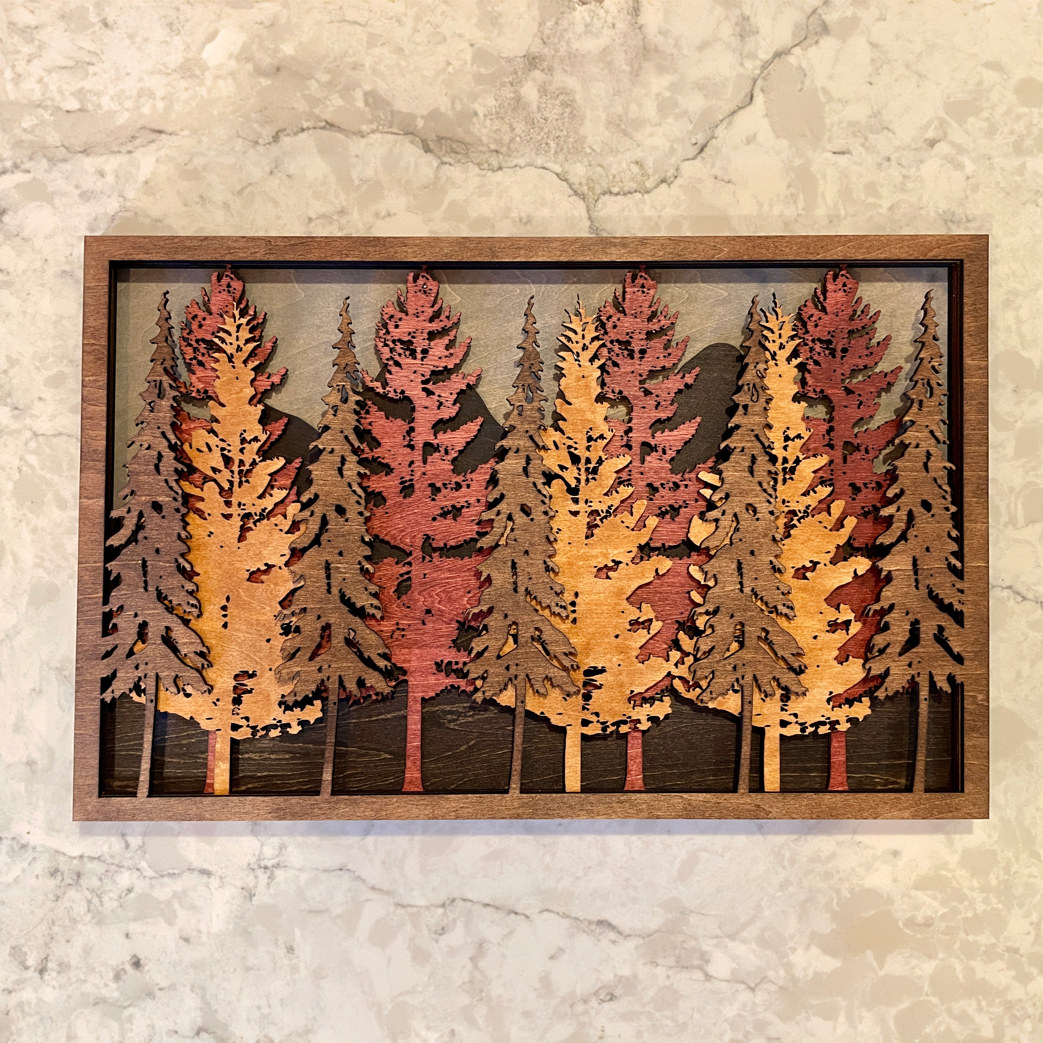3D wood wall art displaying layered mountain peaks and forest trees in various stains, handcrafted by Encinitas 3D, set against a marble background.