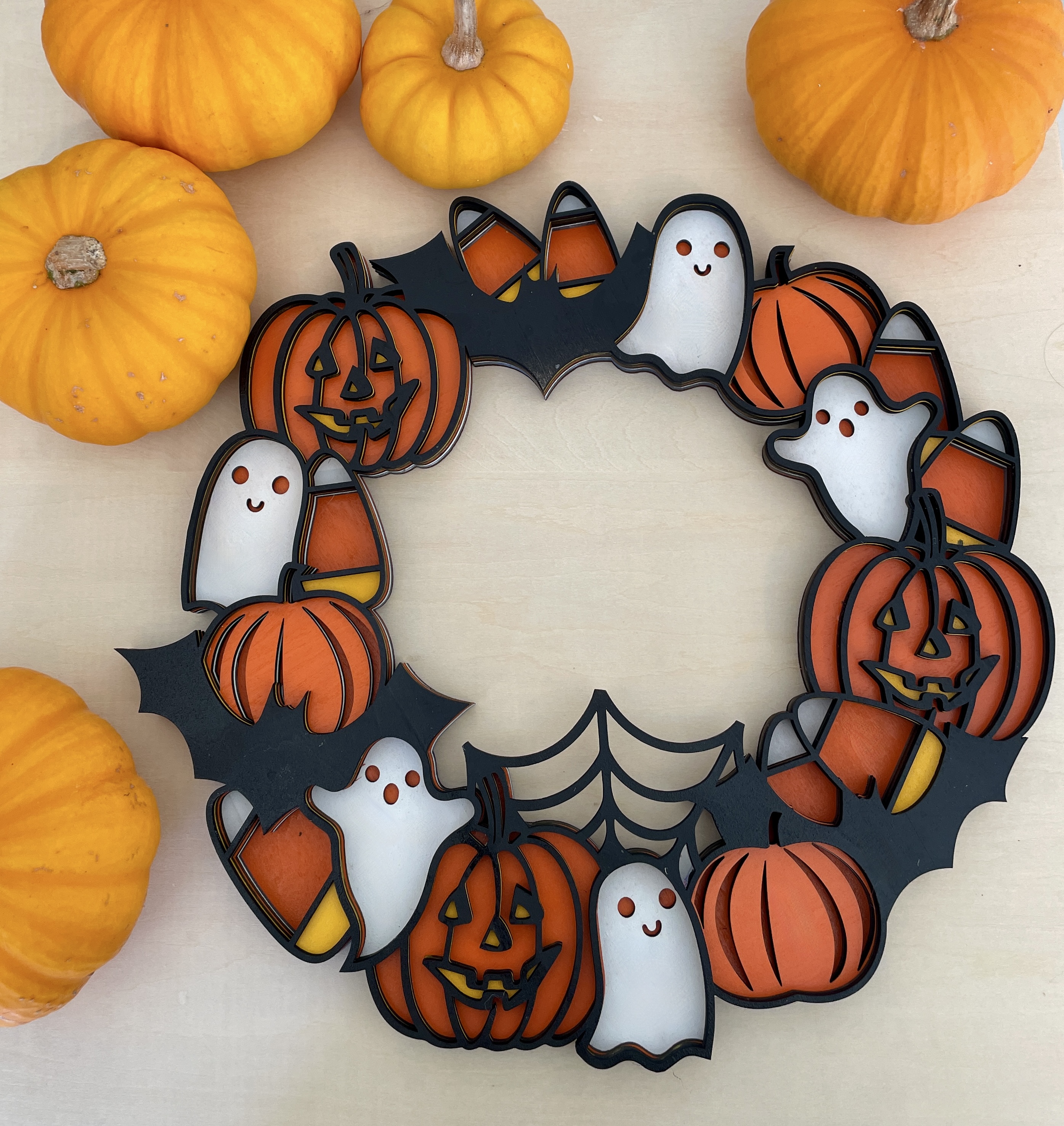 Glow-in-the-dark Halloween wreath with intricate laser-cut layers featuring candy corn, pumpkins, ghosts, bats, and spider webs. Vibrant, hand-painted festive colors. Ideal spooky decor for doors and walls. encinitas 3D encinitas3D.com