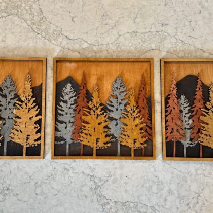 Three-piece 3D wood wall art displaying layered mountain peaks and forest trees in various stains, handcrafted by Encinitas 3D, set against a marble background.