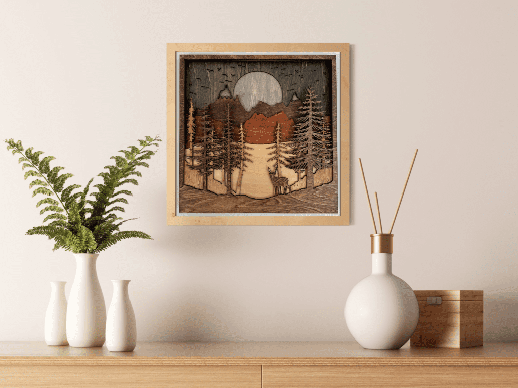 A physical wall art piece featuring a mountain mandala design, composed of seven stained basswood layers, each laser-cut to depict various natural elements. The artwork is 11.5 inches square and 0.75 inches thick, displaying scenes including the moon, birds, mountains, hills, a blue lake, trees, and a deer, all crafted with detailed wood grains to enhance its rustic aesthetic.