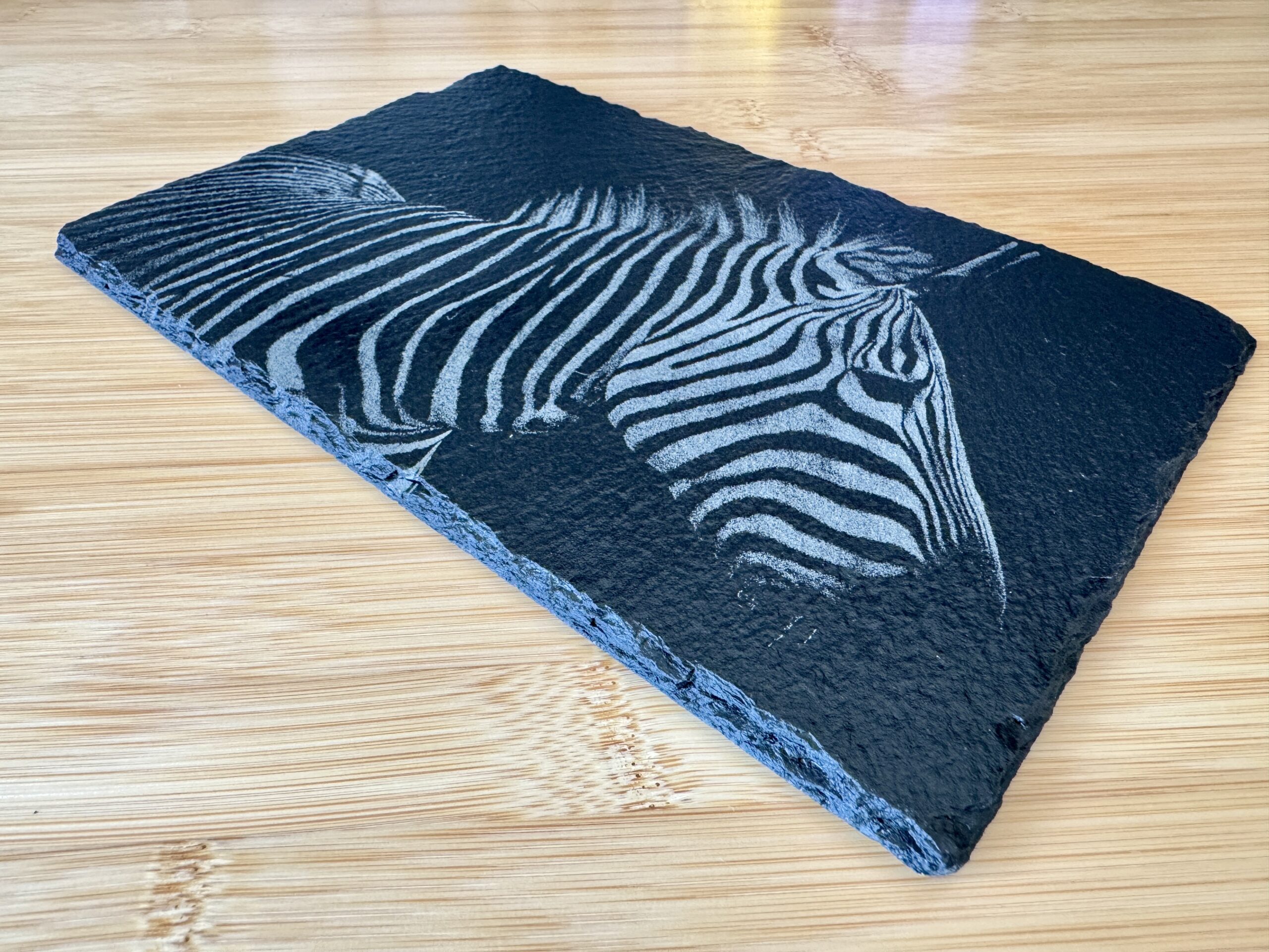 “High-contrast black and white image of a zebra with detailed stripes engraved onto a natural slate tile. The unique texture of the slate adds depth to the zebra’s distinct patterns, creating a striking, artistic effect. Perfect as a decorative piece or a custom gift from Encinitas3D.com, where technology meets nature-inspired design.”
